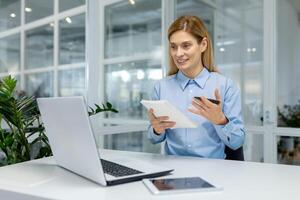un mujer en un moderno oficina espacio es enfocado en aprendizaje y tomando notas mientras trabajando en su digital tableta. foto