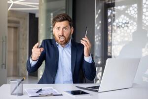 Portrait of an angry young man businessman sitting in the office at the table, talking on the phone, shouting and pointing his finger at the camera accusing someone. photo