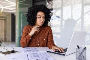 grave pensando mujer trabajador financiero trabajando con documentos y documentos dentro oficina a lugar de trabajo, mujer de negocios comprobación informes, utilizando calculadora y ordenador portátil en papeleo. foto