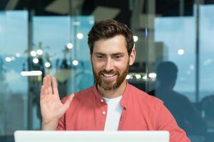 call of successful and cheerful smiling businessman, man talking remotely using laptop while sitting inside office, online meeting with colleagues and helping customers. photo