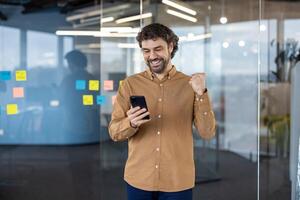 Satisfied millennial male in sandy shirt making victory gesture while looking at screen of mobile phone with happy expression. Lucky guy reading message with good news while going to workplace. photo