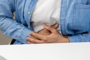 Close-up photo of a body part, hands of a young Latin American man in a blue shirt holding his stomach, feeling and suffering from pain.