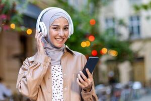 sonriente Arábica mujer en hijab escuchando audio libro en auriculares conectado a móvil dispositivo mientras caminando abajo calle. atractivo dama disfrutando ocio y utilizando moderno tecnología en borroso antecedentes. foto