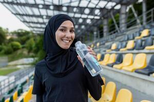 un contento musulmán mujer en Deportes atuendo y hijab participación un agua botella a un Deportes estadio, simbolizando salud y aptitud física. foto