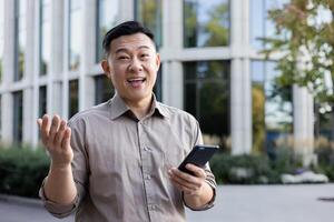 Portrait of a happy young Asian male office worker standing near an office center and holding a phone. photo