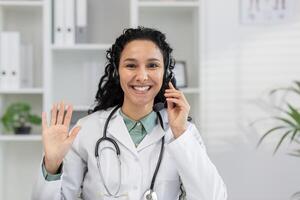 alegre hembra médico vistiendo un auriculares saluda pacientes durante un médico virtual consulta. profesional cuidado de la salud y tecnología concepto representado en un moderno clínica ambiente. foto