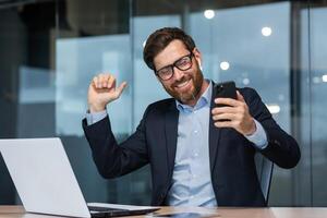 Successful mature businessman in office listening to music and dancing, boss in glasses with phone celebrating achievement working inside building using laptop at work. photo
