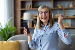 Elderly woman holding a smartphone in a casual indoor setting appears elated or triumphant, possibly celebrating good news. photo