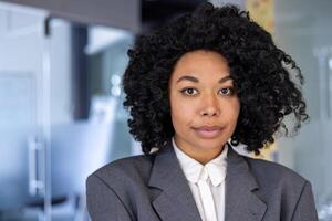 cerca arriba retrato de grave confidente negocio mujer, africano americano mujer con Rizado pelo y en negocio traje mirando a cámara, hembra trabajador dentro oficina a lugar de trabajo. foto