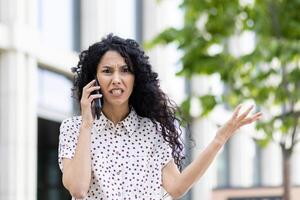 Curly-haired woman in a spotted blouse expressing concern while talking on the mobile phone, an office building backdrop. photo