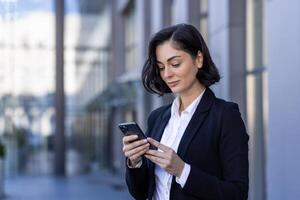 un joven negocio mujer es en pie cerca el oficina centrar en el calle en un negocio traje y utilizando el teléfono, mecanografía mensajes, enviar mensajes de texto foto