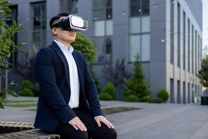 Serious and focused Asian male office worker in a business suit sitting on a bench near a building wearing a virtual mask. photo