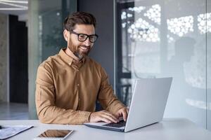 Experienced programmer inside office at workplace, senior man with beard and glasses working on laptop, businessman satisfied with achievement results. photo