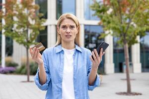 Blue eyed woman standing next to shopping mall with debit card and smartphone in raised hands. Stressed female feeling frustrated because of uncharged device and inability checking bank balance. photo