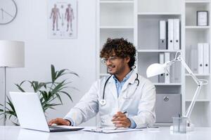 joven médico trabajos con un ordenador portátil dentro el médico oficina de el clínica, el hombre felizmente sonrisas y llena fuera médico informes y tratamiento formularios, trabajos con documentos. foto