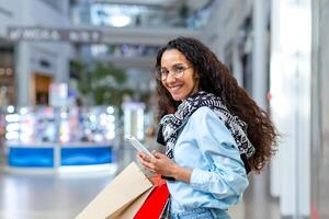 un joven latín americano niña estudiante compras en historias. él es participación un teléfono, paquetes con bienes foto