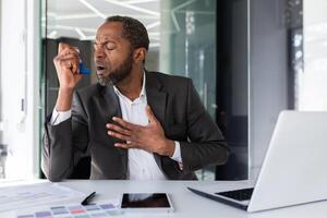 Sick man inside office with asthma using inhaler to ease breathing, mature experienced african american man coughing working sitting with laptop at workplace. photo