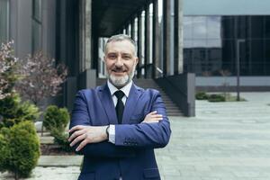 retrato de un exitoso mayor hermoso hombre, empresario, juez, abogado. en pie en un traje cerca el palacio de justicia, oficina centro. cruzado su brazos en frente, mira a el cámara, sonrisas foto