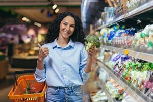 retrato de un hermosa vegetariano mujer en un supermercado, un Hispano mujer sonriente y mirando a el cámara y sonriente elige brócoli vegetales en un tienda de comestibles Tienda foto