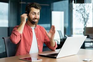 Mature successful businessman with call headset working inside office, man consulting online clients, talking with colleagues on online remote meeting, sitting at workplace with laptop. photo