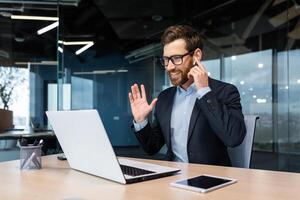 Online meeting with clients, conference. A young man businessman in a suit sits in the office at the table. Talks on a call from a laptop. He holds the earphone in his hand, says hello, smiles. photo