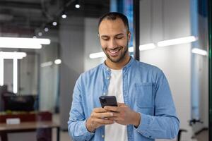 indio Hispano hombre en casual atuendo, contento mientras mirando a su teléfono dentro un contemporáneo oficina ajuste. foto