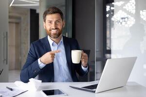 Portrait of a young man working in the office, sitting at a desk in a suit, holding a cup in his hand and pointing at it with his finger, smiling and looking at the camera. photo