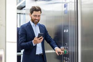 un joven sonriente empresario y abogado es en pie en el ascensor de un oficina edificio y prensado el incorrecto botón mientras utilizando un móvil teléfono. foto