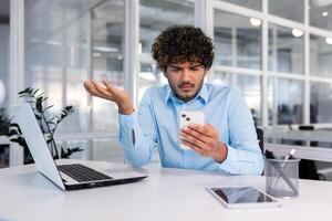 empresario dentro el oficina a el lugar de trabajo es no satisfecho con el trabajo de el teléfono, el hombre es trastornado participación un teléfono inteligente en su manos, Hispano hombre es sentado a el mesa con un ordenador portátil. foto