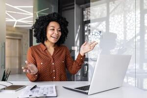 alegre exitoso mujer de negocios hablando remotamente utilizando ordenador portátil para llamar, hembra empleado sonriente y gesticulando mirando a computadora pantalla, trabajando dentro oficina a lugar de trabajo. foto