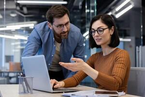 Confident caucasian secretary in eyeglasses showing screen of laptop to attentive bearded head of company. Successful coworkers discussing plans and goals for week while making list of meetings. photo