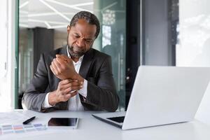 Overtired and overworked man at workplace sitting inside office, businessman has wrist joint pain on his hand, african american man in business suit working long time sitting. photo