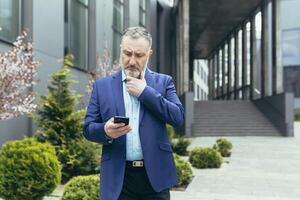Pensive and worried senior man in a business suit stands on the street near a modern building, looks at a mobile phone, reads, types, holds his beard thoughtfully. photo