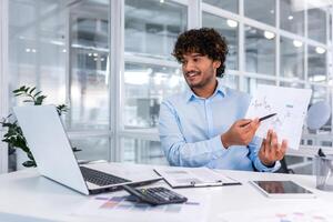 Portrait successful businessman, Latin American man shows a report of financial documents online interlocutors in the laptop screen, an employee inside an office building online conference. photo