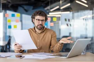 Stressed man wearing sandy shirt to office and stretching hand to portable computer while holding document. Confused guy noticing mistake in report and checking electronic version of calculations. photo