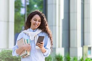 un joven hermosa Hispano mujer camina ciudad con un teléfono en manos, un estudiante con libros fuera de el instalaciones de un Universidad académico biblioteca usos un solicitud en un teléfono inteligente foto