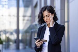 problemas con el financiero cuenta. trastornado joven negocio mujer en pie en el calle y participación su cabeza, participación un crédito tarjeta y mirando a el teléfono. foto