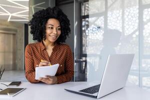 A young professional woman is engaged in a business meeting, taking notes in a modern office setting with natural light and a laptop in front of her. photo