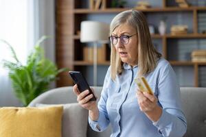 un más viejo mujer mira confuso mientras participación un crédito tarjeta y mirando a su teléfono inteligente, posiblemente relación comercial con en línea compras o tecnología retos foto