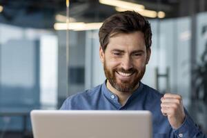 alegre masculino profesional con barba en oficina configuración, sonriente mientras mirando a ordenador portátil pantalla, expresando éxito y satisfacción. foto