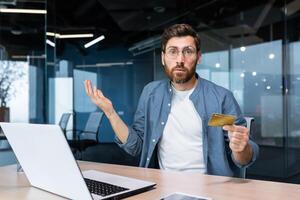 Portrait of disappointed and deceived businessman inside office, mature man looking at camera confused holding bank credit card and phone, employee got rejection error. photo