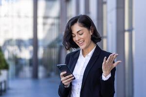 sonriente joven mujer de negocios en pie en el calle cerca un oficina edificio, utilizando el teléfono, contento con bueno noticias, leyendo un mensaje. foto