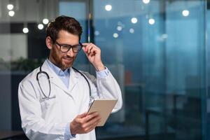 Cheerful and smiling senior doctor in glasses and medical gown reading message from tablet computer, man working inside modern clinic photo