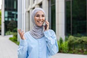 Young joyful businesswoman in hijab talking on the phone outside an office building. photo