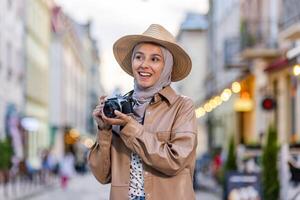 joven hermosa mujer caminando en el noche ciudad en hiyab, turista con cámara y vistiendo un sombrero inspecciona el histórico ciudad sonriente con satisfacción, musulmán mujer en un viaje. foto