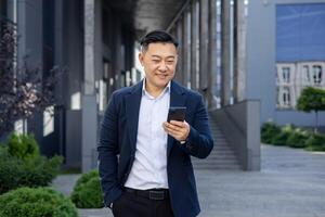 A content Asian businessman in a suit enjoys a sunny day outside modern buildings while browsing his phone. photo