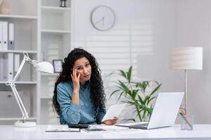 un Hispano mujer de negocios aparece confuso mientras trabajando en su hogar oficina, rodeado por su trabajo esenciales foto