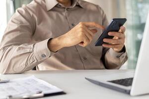 sección en medio de un profesional hombre utilizando un teléfono inteligente mientras sentado a un moderno oficina escritorio con un ordenador portátil. atención en digital comunicación en un negocio ajuste. foto