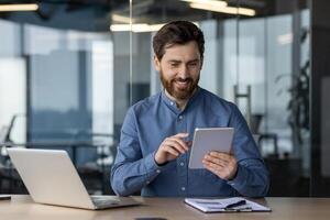 alegre barbado hombre en casual atuendo disfrutando su tableta con un sonrisa a un organizado escritorio en un contemporáneo oficina ambiente. foto