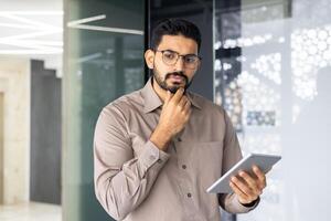 Pensive businessman holding a digital tablet, deep in thought inside a contemporary corporate environment. photo